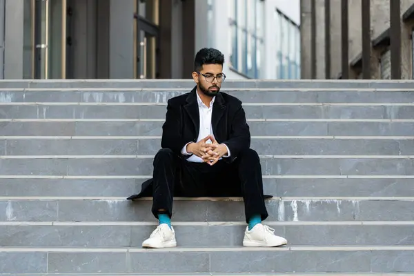 Unhappy sad Indian business man frustrated stress pensive feeling bad after company unemployment lost job problem solution crisis outdoor. Arabian Hindu freelancer guy sitting on stairs downtown city