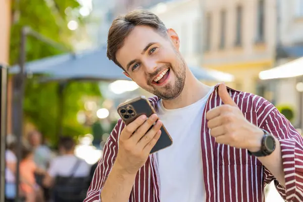 Excited Caucasian man use mobile smartphone celebrating win good message news, success, lottery jackpot victory, giveaway online outdoor. Happy young guy tourist walking on city street. Lifestyles
