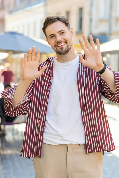 Bearded man smiling friendly at camera, waving hands gesturing invitation hello, hi, greeting, goodbye, welcoming with hospitable expression outdoors. Guy walking in urban city town street. Vertical