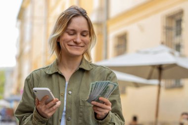 Mutlu genç bir kadın nakit para sayıyor, akıllı telefon hesap makinesi uygulaması kullanıyor, planlanmış tatil hediyeleri için gelirden tatmin oluyor. Şehrin güneşli caddesinde beyaz bir kız. Şehir yaşam tarzları açık havada.