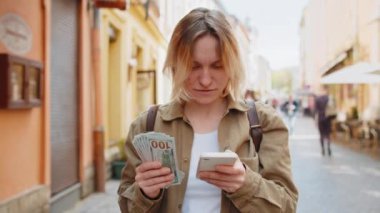 Mutlu genç bir kadın nakit para sayıyor, akıllı telefon hesap makinesi uygulaması kullanıyor, planlanmış tatil hediyeleri için gelirden tatmin oluyor. Şehrin güneşli caddesinde beyaz bir kız. Şehir yaşam tarzları açık havada.