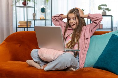 Dizüstü bilgisayar kullanan mutlu kız çocuğu, iyi haber mesajı alıyor, ani zaferle şoke oluyor piyango galibiyetini evde online alışveriş yaparak kutluyor. Yaşam tarzı teknolojileri.