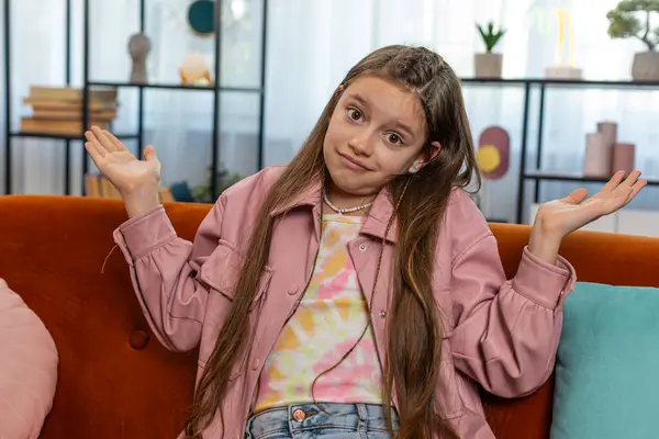 stock image I dont know what to say. Confused young girl child feeling embarrassed about ambiguous question, having doubts, no idea, being clueless and uncertain. Teen kid sitting on couch in living room at home