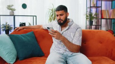 Hüzünlü Arap adam akıllı telefon daktilo kullanıyor, beklenmedik piyango sonuçlarını kaybediyor, kötü haber, servet kaybı, başarısızlık, teslim tarihi, virüs. Genç Hintli adam kanepede oturuyor.