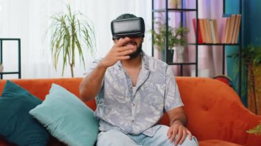Hintli adam sanal gerçeklik teknolojisi VR uygulama kulaklığı kullanarak 3D video oyunu simülasyonu yapıyor ve modern ev dairesinde film izliyor. Kanepede oturan gözlüklü adam.