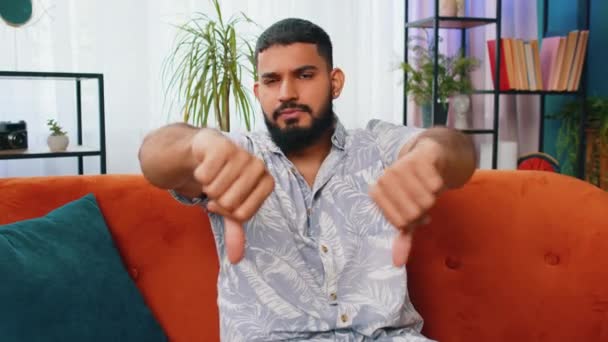 気に入らない 不幸な若いアラビア人男性は サインジェスチャーの親指を示し 不満を表明し 家のアパートで不満の悪い仕事を屋内で表現しました ソファーの部屋で不満なインディアン男 — ストック動画