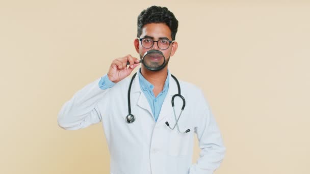 快乐的印度年轻医生心脏病专家男人举着放大镜 牙齿上挂着滑稽的笑脸 傻笑着 口腔科米色背景的阿拉伯药剂师 — 图库视频影像