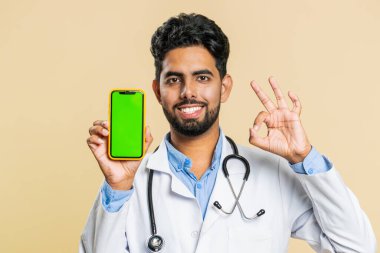 Hintli genç doktor kardiyolog elinde yeşil ekran krom anahtarı olan akıllı telefon ile iyi bir uygulama tanıtım teklifi sunuyor. Tamam, telefon numarası. Bej arka planda Arap eczacı