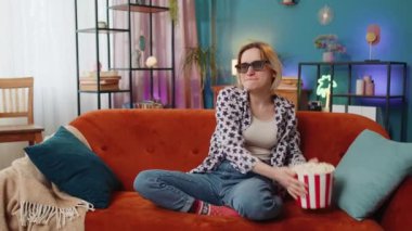 Kanepede oturup patlamış mısır yiyen ve ilginç TV dizileri, spor oyunları, filmler, evdeki çevrimiçi sosyal medya filmleri izleyen heyecanlı bir kadın. Üç boyutlu gözlüklü kız ev eğlencesi yapıyor.