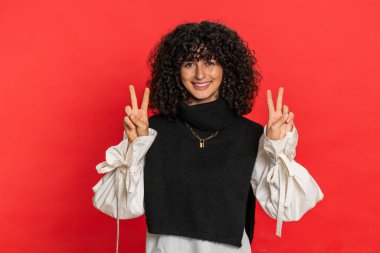 Zafer işareti gösteren kıvırcık saçlı mutlu beyaz kadın, başarı ve kazanma umuduyla barış jesti yapıyor, iyimser bir ifadeyle gülümsüyor, kutluyor. Kırmızı arkaplanda izole edilmiş kız