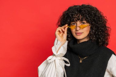 Kıvırcık saçlı, turuncu güneş gözlüğü takan, şık gülümseyen beyaz kadın kameraya bakıp hayal kuran, dinlenen, iyi haberler veren mutlu bir yüz ifadesi. Kırmızı arkaplanda izole edilmiş kız