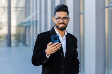 Mutlu Hintli işadamı akıllı telefon yazıcı kullanıyor e-posta mesajlarına cevap veriyor açık havada online mobil uygulamalarla sohbet ediyor. Şehir merkezindeki kameraya bakan Arap Hintli adam. İş adamları