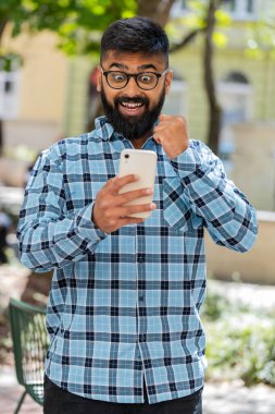 Hintli sakallı adam cep telefonuyla kutlama yapıyor. İyi haber kazan, piyango büyük ikramiyesi kazan, açık havada dağıtım yap. Şehir sokağındaki İspanyol adam. Kasaba yaşam tarzları. Dikey