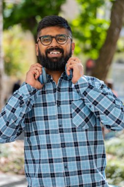 Mutlu, gülümseyen, müzik dinleyen, kulaklık takan, kameraya bakıp rahatlayan Hintli sakallı bir adamın portresi. Şehir sokağındaki Latin kökenli genç adam. Dikey