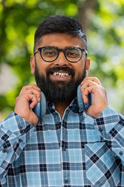 Mutlu, gülümseyen, müzik dinleyen, kulaklık takan, kameraya bakıp rahatlayan Hintli sakallı bir adamın portresi. Şehir sokağındaki Latin kökenli genç adam. Dikey