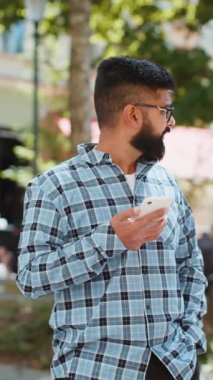 Hintli sakallı adam akıllı telefon kullanıyor sosyal medya uygulamasında mesaj yazıyor, internette sörf yapıyor, dinleniyor, dışarıda mola veriyor. Şehir caddesinde duran İspanyol adam. Dikey