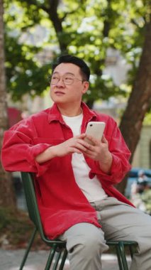 Asyalı orta yaşlı bir adam sosyal medya uygulamasında akıllı telefon kullanıyor, internette sörf yapıyor, dinleniyor, dışarıda mola veriyor. Şehir caddesinde oturan Çinli adam. Dikey