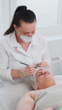 Güzellik uzmanı, genç yetişkin bir kadının alnına fırçayla nemlendirici kimyasal krem maske takıyor. Cilt bakımı prosedürü. Yüz kozmetolojisi tedavisi. Enzim terapisi. Dikey