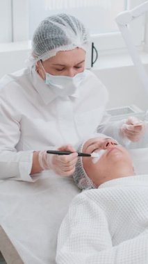Güzellik uzmanı, genç bir kadının yüzüne fırçayla nemlendirici kimyasal krem maske takıyor. Kaplıcadaki kız cilt bakımı. Yüz kozmetolojisi tedavisi. Enzim terapisi. Dikey