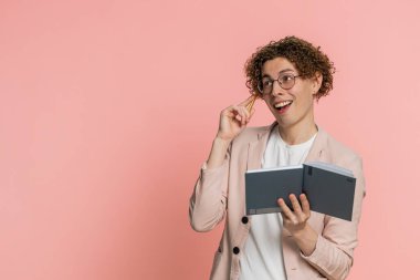 Düşünceli, beyaz, kıvırcık saçlı, ceketli bir gazeteci not defterine not defteri günlüğüyle düşünceler yazıyor, iyi fikir. İçerideki pembe arka planda izole edilmiş genç adam.