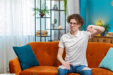 Hoşlanmamak. Mutsuz, genç, beyaz bir adam işaret dilini gösteriyor. Memnuniyetsizliğini dile getiriyor. Memnuniyetsizliğini dile getiriyor. Kanepede kıvırcık saçlı hoşnutsuz adam..