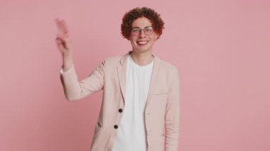 Zafer işareti gösteren kıvırcık saçlı mutlu beyaz adam, başarı ve kazanma umuduyla barış jesti yapıyor, iyimser bir ifadeyle gülümsüyor, kutluyor. Pembe arka planda izole edilmiş adam