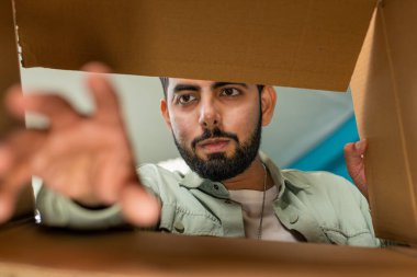 Kutunun içinden bak. Evde paketleri açan mutlu Hintli adam. Gülümseyen memnun erkek alışverişci, çevrimiçi dükkan müşterisi karton kutu açıyor hızlı posta yoluyla satın alma hediyesi alıyor