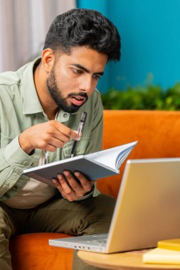 Evde dizüstü bilgisayarla kitap okuyarak serbest çalışan Hintli bir öğrenci. Adam web kamerasıyla konferans görüntülü konuşma yapıyor. Çevrimiçi öğretmen uzaktan sohbette e-öğreniyor. Çevrimiçi uzaktan eğitim. Dikey