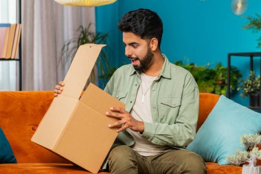 Paketleri açan mutlu Hintli genç adam evde oturuyor. Gülümseyen sakallı Arap adam çevrimiçi dükkan müşterisi karton kutu açan müşteri hızlı posta yoluyla satın alma hediyesi alıyor.