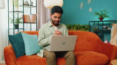 Laptop bilgisayar kullanan Hintli genç adam, iyi haber mesajı alıyor. Aniden kazanılan piyango galibiyetini kutluyor. Evdeki kanepede online alışveriş oyunu oynuyor.