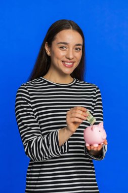 Çizgili bluz giyen mutlu zengin genç kadın gelecek ihtiyaçlar için kumbaraya dolar harçlığı koyuyor. Bozuk parası olan kız gelir artışını finanse ediyor maaş geliri, mavi arka planda ekonomi