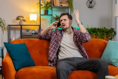 Kızgın genç adam evdeki kanepede otururken akıllı telefonda tartışıyor. Tartışma. Kafkasyalı adam yüksek sesle konuşuyor ve cep telefonuna bağırıyor, kötü niyetle hareket ediyor ve işaret ediyor..