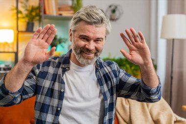 Beyaz, olgun bir adam kameraya dostça gülümsüyor ve iki elini sallayarak merhaba diyor. Evinde misafirperver bir yüz ifadesiyle karşılıyor. Yaşlı sakallı adam evdeki oturma odasında kanepede oturuyor.
