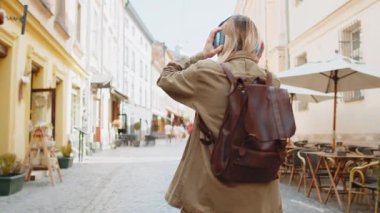 Sırt çantalı genç bir turist sokakta yürürken kulaklıkla müzik dinliyor, etrafa bakıyor ve gülümsüyor. Şehir geçmişindeki kız gezginin arka planı. Yaşam Tarzları.