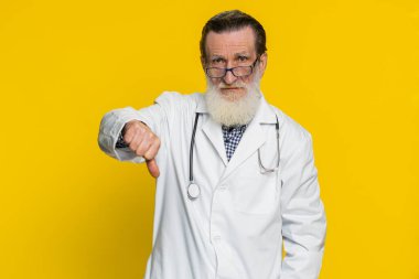 Hoşlanmamak. Mutsuz yaşlı doktor kardiyolog. Başparmak işareti gösteriyor. Onaylanmamış olumsuz geri bildirim. Eczacı dedesi sarı arka planda izole edilmiş.