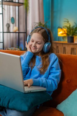 Laptop kullanan, kulaklıklı beyaz ergen bir kız müzik dinliyor ya da ders dinliyor, uzaktan öğreniyor. Online eğitim. Gülümseyen küçük bir çocuk, dinlenen bir çocuk, iş yerinde kanepede oturuyor..