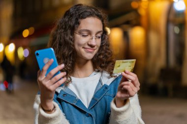 Genç bir kız kredi kartı akıllı telefonu kullanıyor para transfer ederken, online alışveriş yaparken, yemek siparişi verirken, otel odasında rezervasyon yaparken. Şehir caddesinde gülen mutlu bir kadın.