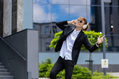 Kablosuz kulaklıklar takmış, dans eden, açık havada dans eden enerjik disko müziği dinleyen mutlu, Kafkasyalı genç iş adamı. Şehir merkezindeki ofis binasının dışında takım elbiseli neşeli bir adam..