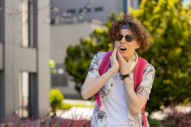 Şehir caddesinde diş ağrısından muzdarip beyaz bir genç turist. Kıvırcık saçlı ve sırt çantalı üzgün erkek elini yanağının yakınında tutuyor, hassas noktasına masaj yapıyor, acı hissediyor. Diş tedavisine ihtiyacı var.