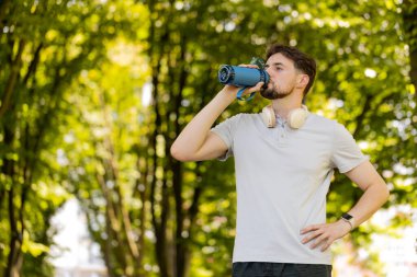 Sportif atletik susamış beyaz erkek koşucu parkta çalıştıktan sonra su içiyor, spor şişesi içiyor aktif genç yaşam tarzı ferahlatıcı su içiyor gülümseyerek uzaklaşıyor..