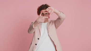 Kafkasyalı fotoğrafçı elleriyle resim çerçevesi çiziyor, parmaklarından bakıyor ve ilginç bir ana odaklanıyor, zoom 'u taklit ediyor ve güzel görüntüler kesiyor. Pembe arka planda izole edilmiş genç adam