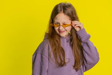 Turuncu güneş gözlüğü takan, gülümseyen, göz kırpan, flört eden, iyimser bir şekilde kameraya bakan çocukluk çağındaki çocuk portresi. Sarı arka planda genç çocuklar