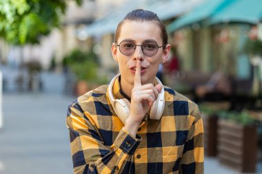 Sessiz olun lütfen. Genç adam işaret parmağını dudaklarına bastırıyor. Sessiz ol işareti yapıyor. Dışarıdaki dedikoduları kimseye anlatmıyor. Şehir caddesinde duran turist adam. Kasaba yaşam tarzları.
