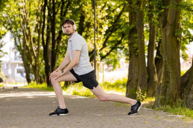 Spor giyim bölümünde spor giyinen genç bir adam hamleler yapıyor ve yaz parkında bacaklarını esnetiyor. Koşu antrenmanından önce ısın. Atletik erkek koşucu ağaçların arasında yaya yolunda bacak çalıştırıyor. Çömelme eğitimi aldı.