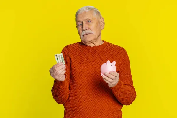 stock image Poor senior old man insufficient amount of money, holding piggybank and one dollar banknote. Financial crisis. Bankruptcy. Poverty and destitution. Elderly grandfather pensioner on yellow background