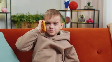 Telefon, iyi haber, dedikodu. Akıllı telefondan hoş sohbetler yapan, çocukluk çağındaki mutlu çocuk arkadaşıyla konuşmaktan zevk alıyor. Ergen, beyaz, erkek, küçük çocuk. Oturma odasında, kanepede.