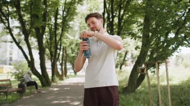 Sportif atletik susamış beyaz erkek koşucu parkta çalıştıktan sonra su içiyor, spor şişesi içiyor aktif genç yaşam tarzı ferahlatıcı su içiyor gülümseyerek uzaklaşıyor..