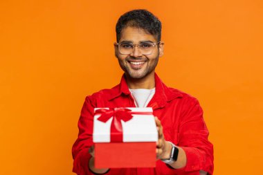 Pozitif gülümseyen Hintli adam doğum günü hediyelerini sunar ellerini uzatır, hediye paketi hediye ikramiyesi teklif eder, kutlama partisi düzenler, indirim indirimi yapar. Turuncu arka planda izole edilmiş Arap adam.