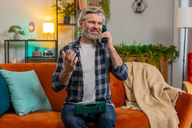 Oturma odasındaki koltukta oturup telefonla konuşan mutlu Kafkasyalı yetişkin bir adam. Heyecanlı sakallı yaşlı adam eski moda telefondan keyif alıyor..