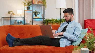Rahat koltukta uzanan uygulamaları kullanarak dizüstü bilgisayarda yazan gülen Hintli bir adam. Evdeki bilgisayarda çalışan mutlu İspanyol genç adam. Netbook 'ta e-öğrenme, internette gezinme